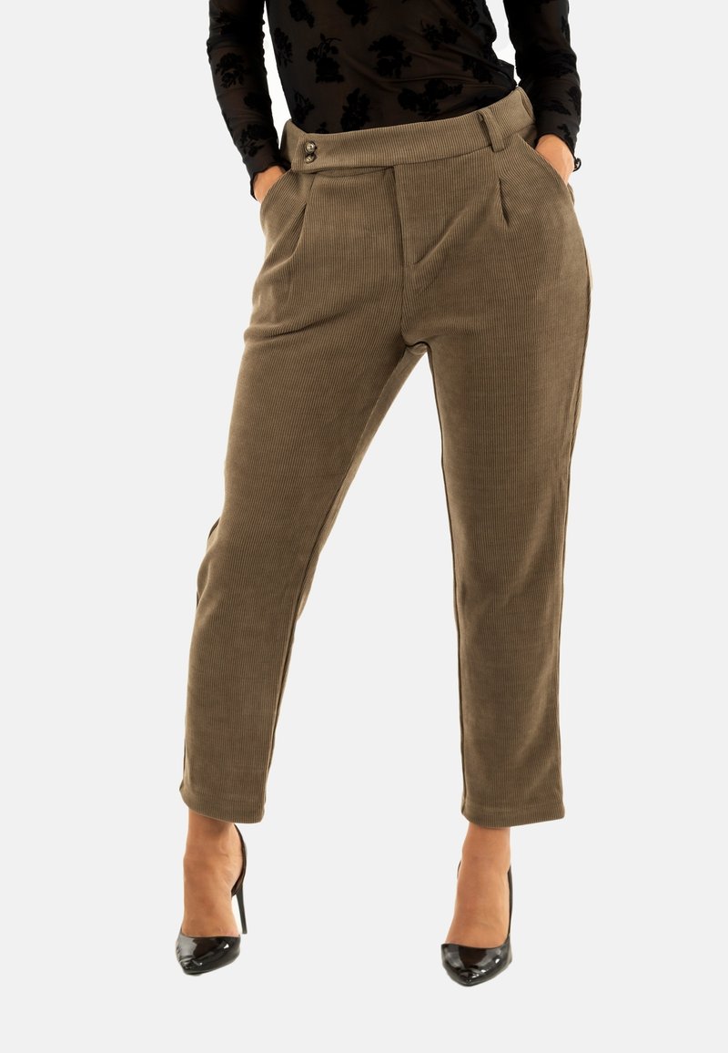 Donna che indossa pantaloni in velluto a coste marroni a vita alta e gamba affusolata, con tasche laterali e tacchi alti neri, con le mani in tasca, in piedi contro uno sfondo bianco.