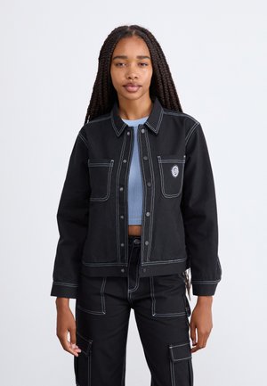 Veste en denim noire avec des coutures blanches contrastantes, fermeture à boutons-pression à l'avant et deux poches poitrine. Portée par-dessus un haut bleu clair.