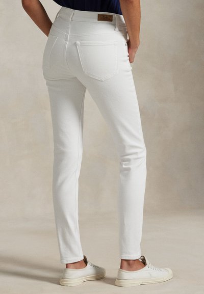 Osoba w białych, dopasowanych dżinsach i białych trampkach, stojąca z rękami opartymi na biodrach na jednolitym tle.