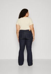 Jeans flare azul-escuro com uma cintura alta, apresentando bolsos traseiros com costura e um remendo bege na cintura. Combinados com uma blusa clara de malha canelada.