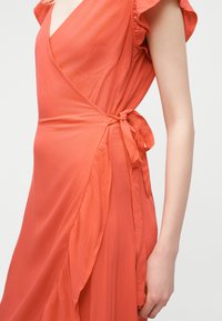 Kvinne iført en korallfarget omslagskjole med flagrende ermer, bundet i midjen, vist fra skulder til midt på låret mot en ensfarget bakgrunn.