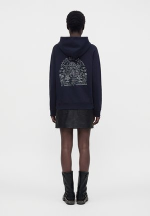 Personne portant un sweat à capuche bleu marine avec le texte « Black Reality I Want Magic » et un motif floral dans le dos, une jupe noire et des bottes noires mi-mollet.