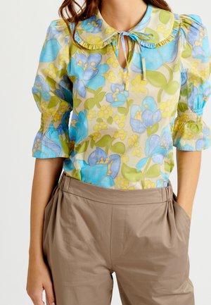 Kobieta w kwiecistej bluzce z bufkami na rękawach, falbanianym kołnierzem z wiązaniem oraz beżowych plisowanych spodniach z wysokim stanem, z rękami w kieszeniach.