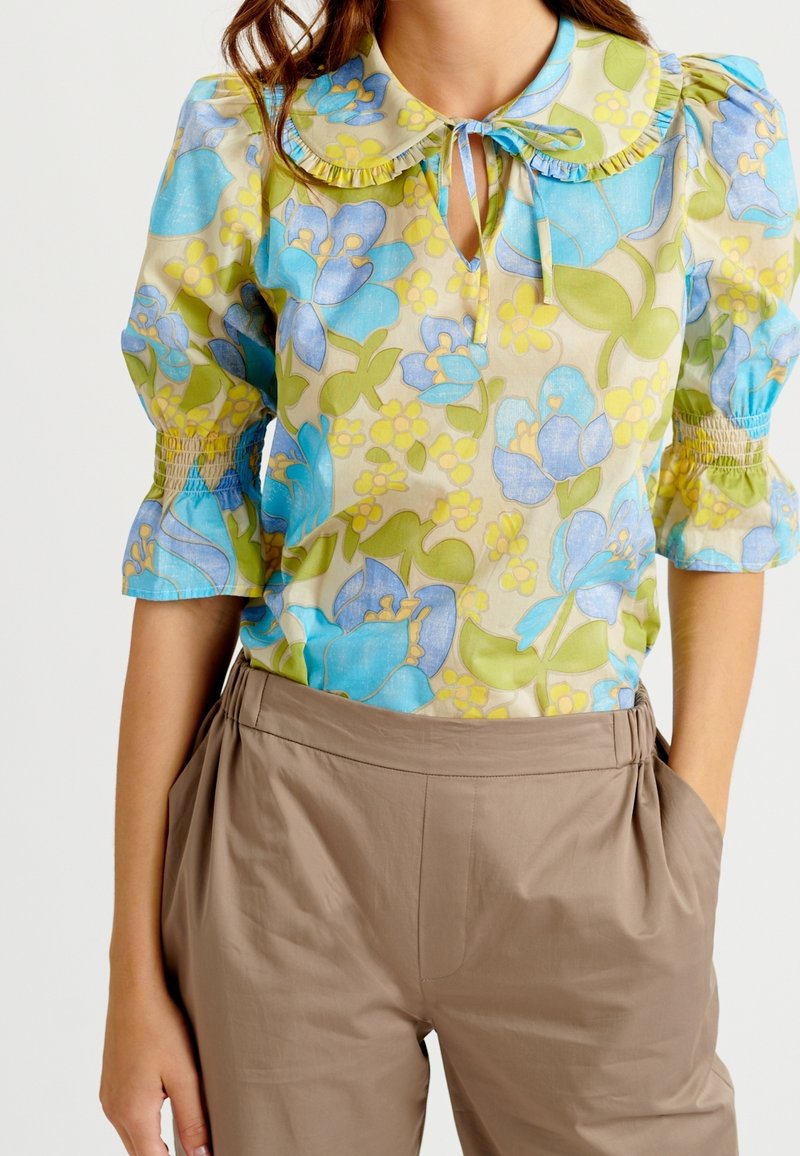 Femme portant un chemisier à fleurs avec des manches bouffantes, un col à volants avec un lien, et un pantalon beige taille haute à plis, les mains dans les poches.