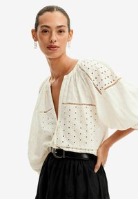Blusa bianca ricamata con maniche a sbuffo, caratterizzata da motivi ergonomici e scollatura a V, abbinata a una gonna nera e una cintura.