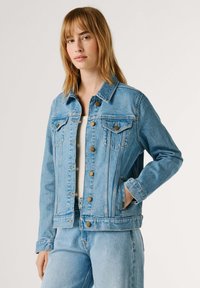 Kvinde med lyst brunt hår iført lyseblå denimjakke og matchende jeans, stående med den ene hånd i jakkelommen mod en ensfarvet baggrund.