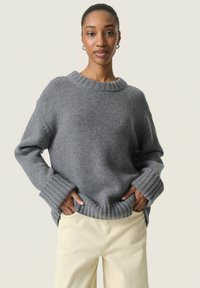Pull en maille gris avec un col et des poignets côtel�és, coupe décontractée, fentes sur les côtés et tissu texturé. Associé à un pantalon de couleur claire.