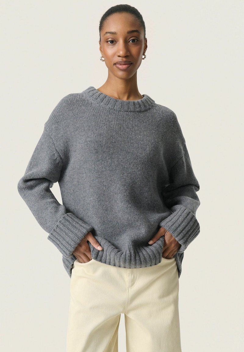 Pull en maille gris avec un col et des poignets côtelés, coupe décontractée, fentes sur les côtés et tissu texturé. Associé à un pantalon de couleur claire.