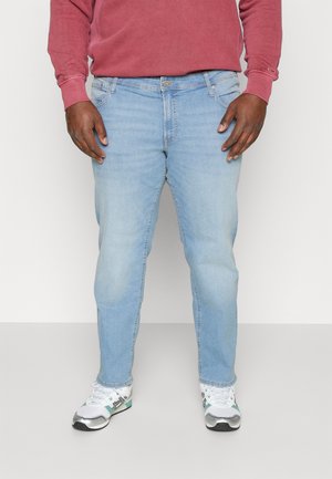 Homme portant un jean bleu clair, des baskets blanches et un sweat-shirt rouge à manches longues, debout devant un fond clair uni.