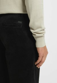 Czarne spodnie z koturnowego materiału z żebrowaną teksturą, w zestawieniu z jasnobeżowym swetrem. Posiadają tylną kieszeń z metką marki.