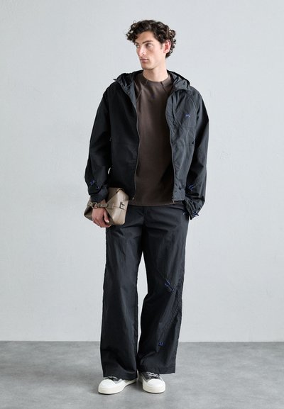 Black lightweight jacket with a hood and blue accents, paired with loose black pants and a brown top. Model holds a beige bag. White sneakers.
