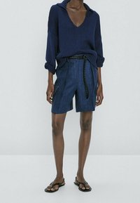Marineblauer Strickpullover mit V-Ausschnitt, kombiniert mit Jeansshorts, die einen geflochtenen Gürtel und seitliche Reißverschlusstaschen haben. Das Model trägt Flip-Flops.