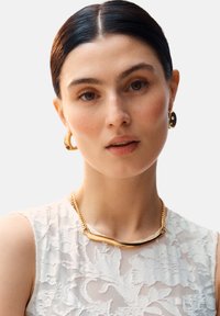 Guldtonad halsband med en slät, böjd design; kedjelänkar; parat med runda guldörhängen med svart inlägg. Modell iförd en spetsblus.