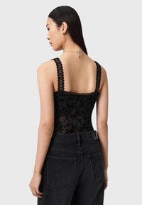 Schwarzer Spitzen-Body mit Blumenmuster und gezackten Trägern, kombiniert mit hoch taillierten schwarzen Jeans. Haare glatt und seidig gestylt.