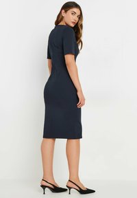 Marineblå tætsiddende kjole med korte ærmer, knælængde, diskrete sømdetaljer og høj halsudskæring. Sammen med sorte slingback-støvler.