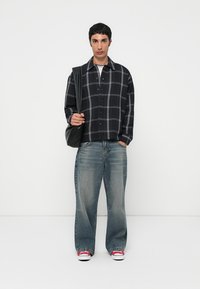 Camisa de cuadros negros con líneas blancas, combinada con jeans de pierna ancha azul claro y zapatillas rojas. El modelo sostiene un bolso negro.
