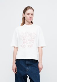 Žena nosi bijelu Carhartt majicu s grafikom lava, uparenom s visokim plavim trapericama, stojeći protiv jednostavne bijele pozadine.