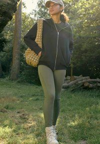 Crna zip-up dukserica, maslinaste zelene tajice, pletena bež torba, bijeli tenisice s čarapama do gležnjeva, stoji na travi s drvećem u pozadini.