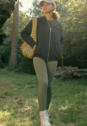 Crna zip-up dukserica, maslinaste zelene tajice, pletena bež torba, bijeli tenisice s čarapama do gležnjeva, stoji na travi s drvećem u pozadini.
