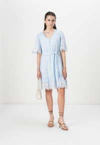 Abito in cotone azzurro chiaro con motivi a occhiello, maniche a campana e orlo scallop. Abbinato a sandali nudi con cinturino e una piccola borsa bianca.