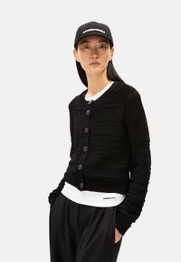 Schwarze Strickjacke mit strukturierten Mustern und sechs Knöpfen, übereinander getragen über einem weißen Hemd. Das Modell trägt eine schwarze Mütze mit weißer Schrift.