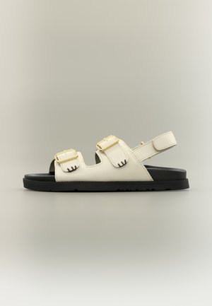 Sandale blanche à bout ouvert avec deux boucles dorées et une bride arrière ajustable, posée sur une semelle noire sur un fond gris uni.