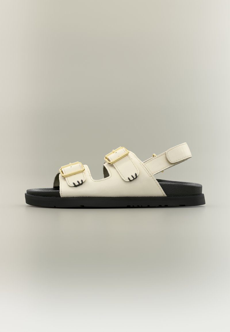 Λευκό σανδάλι ανοιχτού δαχτύλου με δύο χρυσά αγκράφες και ρυθμιζόμενο πίσω λουράκι, τοποθετημένο σε μαύρη σόλα πάνω σε απλό γκρι φόντο.