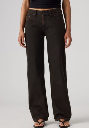 Person iført højttaljede mørkebrune vidde jeans, sort kort top og sorte thongsandaler, der står mod en ensfarvet baggrund.