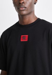 T-shirt en coton noir avec un patch rouge carré portant le logo de la marque. Encolure ronde et manches courtes, texture lisse.