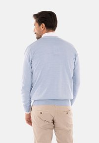 Maglione azzurro chiaro con una texture morbida e orlo a coste, indossato sopra una camicia bianca con colletto, abbinato a pantaloni beige.
