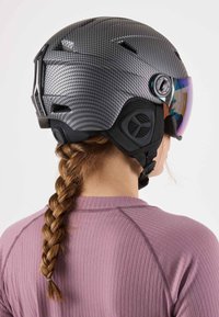 Black carbon-fiber ski helmet with a glossy finish, integrated visor, adjustable strap, and ventilation holes, viewed from the back.