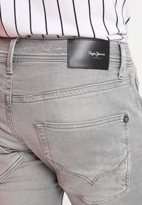 Jeans in denim grigio presentano una texture liscia, tasche standard e un'etichetta nera "Pepe Jeans London" sulla vita.