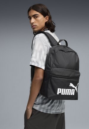 Joven con cabello largo y oscuro lleva una mochila negra Puma, viste una camisa gris con estampado y pantalones oscuros sobre un fondo liso gris.