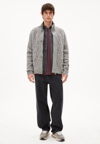 Grauer gestrickter Cardigan mit Rautenmuster, Knopfverschluss, getragen über einem dunklen Hemd und einem weinroten Oberteil, kombiniert mit schwarzen Hosen und grauen Turnschuhen.