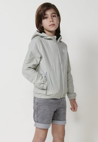 Giacca a vento verde chiaro con zip frontale, polsini elasticizzati e logo. Abbinata a pantaloni corti grigi con orli arrotolati.