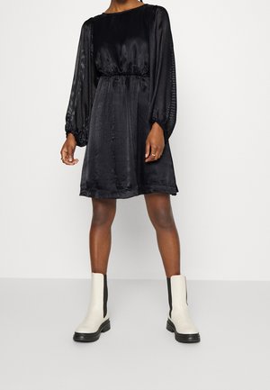 Femme portant une robe noire à manches longues avec une taille cintrée et des bottines blanches, debout devant un fonds clair uni.
