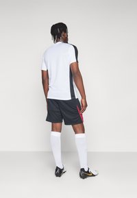 Herrsportkläder med en vit kortärmad skjorta med svarta detaljer, svarta shorts med en röd detalj och vita knästrumpor.