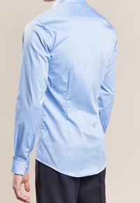 Man som bär en figurnära ljusblå skjorta och mörka byxor, sedd bakifrån mot en beige bakgrund.