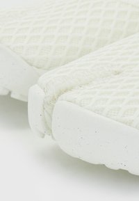 Lightweight white sneaker featuring a textured mesh upper and a solid, speckled rubber sole. Prominent stitching detail along the edges.