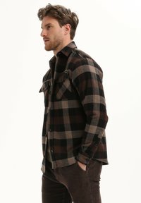 Bruin en zwart geruite flanellen shirt met twee borstzakken en knoopsluitingen, gedragen over donkerbruine broek. Zachte textuur, klassiek kraagontwerp.
