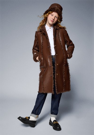 Brown leather coat with fur trim, button closure, and side pockets. Paired with dark denim jeans and black loafers with gold lettering.