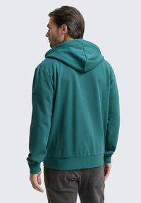 Mann, der von hinten schaut, trägt ein türkisfarbenes Kapuzensweatshirt und dunkelgraue Jeans vor einem einfarbigen hellen Hintergrund.