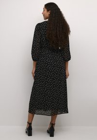 Vestido largo negro con lunares, fabricado en tejido transparente, con cintura elástica y mangas abullonadas. Unos tacones negros completan el conjunto.