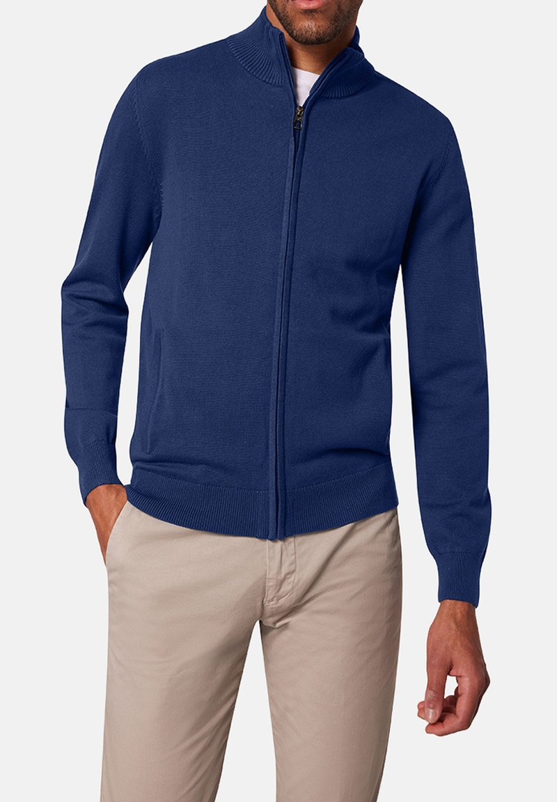 Cardigan zippé bleu marine en tissu tricoté lisse, avec un col montant et des poignets et ourlets côtelés. Comprend deux poches latérales et une fermeture éclair frontale.