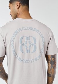 Bărbat cu brațe tatuate purtând un tricou bej deschis, având un logo circular "Closure London Studios" pe spate.