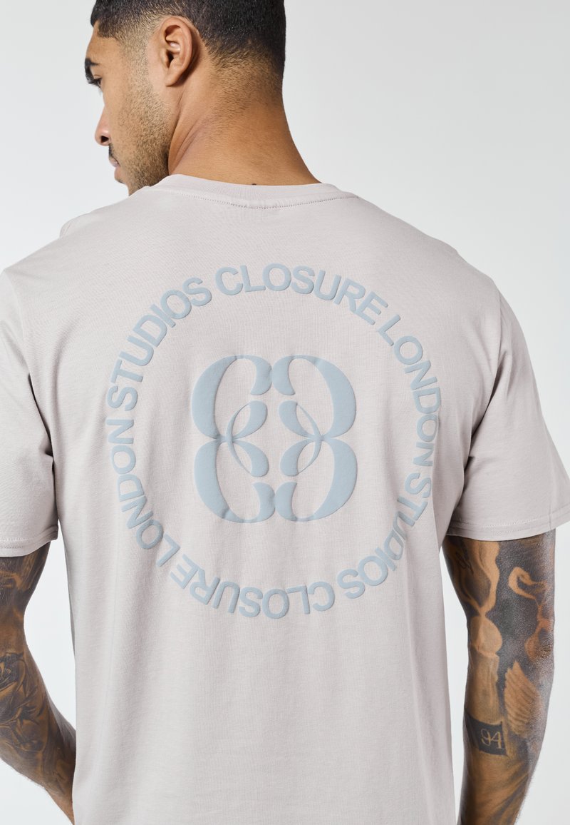 Bărbat cu brațe tatuate purtând un tricou bej deschis, având un logo circular "Closure London Studios" pe spate.