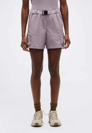 Person iført lyse lilla cargoshorts med bælte, brune sokker og beige sneakers, stående mod en ensformig lys baggrund.