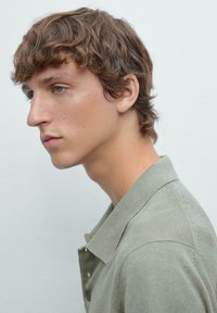 Profilo di una persona con capelli ricci e castani che indossa una camicia color verde chiaro con colletto. Il tessuto ha una consistenza morbida e dettagli di cucitura visibili.