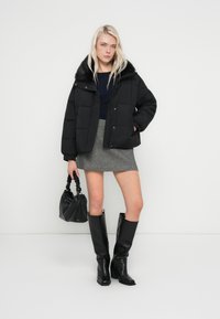 Crna puhasta jakna s ovratnikom od umjetnog krzna, siva mini suknja, crne čizme do koljena i crna torba s pletenom drškom.