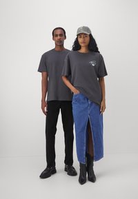 Dos personas están de pie una al lado de la otra. La de la izquierda lleva una camiseta gris y pantalones negros; la de la derecha lleva una camiseta gris, una falda de mezclilla azul y botas negras.